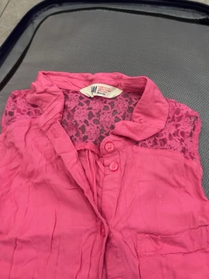 Rosa ärmlös blus med spets från H&M - Snygg rosa blus från H&M med spetsdetaljer upptill och knappar framtill. Blusen är ärmlös och har en klassisk krage samt en bröstficka. Perfekt för en trendig och färgglad look.