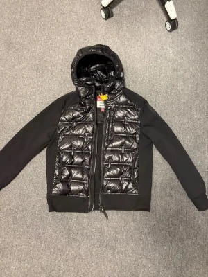 Svart dunjacka med huva Parajumpers - Snygg svart dunjacka från Parajumpers med glansig, quiltad front och huva. Jackan har släta svarta ärmar och ribbade muddar. Dragkedja framtill och diskret Parajumpers-logga på bröstet. Perfekt för kalla vinterdagar.