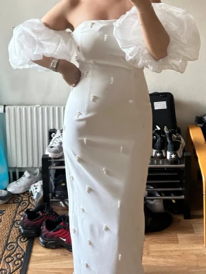 Vit långklänning - Säljer min förlovnings klänning. En vit långklänning med stora puffiga offshoulder-ärmar i transparent tyg. Klänningen har en figurnära passform och små pärlor som detaljer över hela tyget. Perfekt för dig som vill sticka ut med en unik och trendig look.