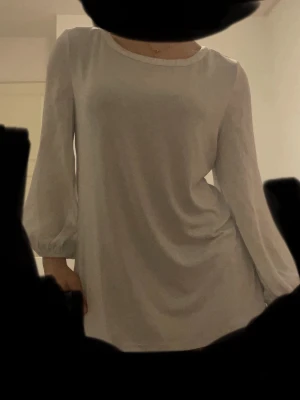 Vit långärmad blus i chiffong - Stilren vit blus med rund halsringning och långa, lätt puffiga ärmar. Tillverkad i ett tunt, flytande chiffongliknande material som ger ett elegant fall. Perfekt att matcha med jeans eller kjol för en clean look.