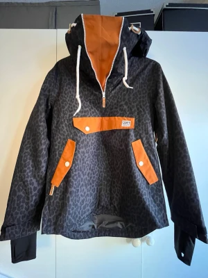 Leopardmönstrad parkas från CWA - Svart parkasjacka med leopardmönster och orange detaljer från CWA. Jackan har stor huva med dragsko, halvlång dragkedja framtill och stora fickor med orange lock och vita knappar. Materialet är troligtvis vattenavvisande syntet. Perfekt för dig som gillar streetstyle och vill sticka ut.