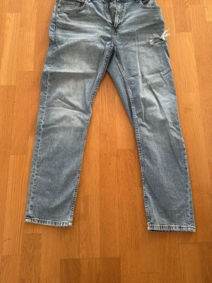 Blå  jeans med slitning - Säljer ett par ljusblå mom jeans med hög midja och avslappnad passform. Jeansen har en snygg slitning på höger lår och klassiska fem fickor. Tillverkade i denim med dragkedja och knapp framtill.
