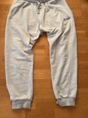 Ljusgrå mjukisbyxor EET CARTEL - Snygga ljusgrå mjukisbyxor från EET CARTEL med resår i midjan och dragsko. Byxorna har ribbade muddar vid bensluten och en enkel, clean design med liten logga på ena benet. Perfekta för chill dagar eller när du vill ha det bekvämt.