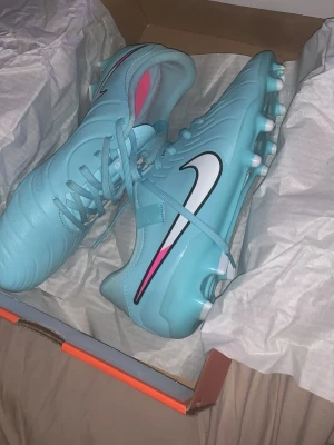 Nike blåa fotbollsskor med dobbar - Snygga ljusblå fotbollsskor från Nike med vita och svarta detaljer samt rosa inslag. Skorna har klassisk snörning och slitstarka dobbar för bra grepp på planen. Tillverkade i syntetmaterial för lätt känsla och bra bollkontroll.