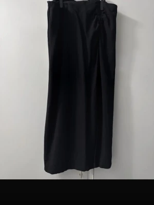 Svart midikjol med slits Small - Stilren svart långkjol i omlottmodell med slits framtill. Kjolen har rynkad detalj i midjan och ett mjukt, lätt tyg som ger ett snyggt fall. Perfekt för dig som gillar minimalistisk och tidlös stil.