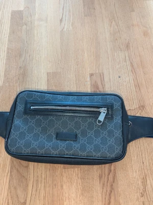 Gucci axelväska med GG-mönster - Snygg axelväska från Gucci i grått med klassiskt GG-mönster över hela väskan. Framsidan har en silverfärgad dragkedja och detaljer i svart läder. Axelremmen är justerbar och har röd-blå rand. Materialet är canvas med läderdetaljer och mesh på baksidan.