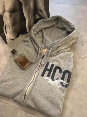 Grå zip-hoodie från Hollister - Snygg grå hoodie med dragkedja från Hollister. Stor HCO-logga och vit mås på bröstet samt texten CALIFORNIA längs ena ärmen. Mjuk bomullsblandning, två fickor fram och klassisk huva. Perfekt för en avslappnad och sportig stil.