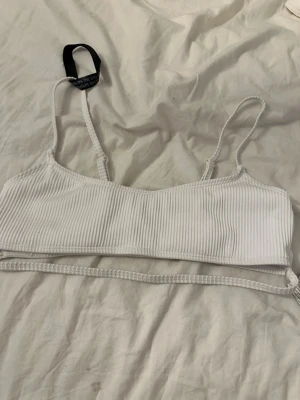 Vit ribbad bikiniöverdel med snörning - Säljer en stilren vit bikiniöverdel med tunna axelband och ribbad struktur. Modellen har en extra snörning nedtill som kan knytas runt midjan för en trendig look. Perfekt för dig som gillar minimalistisk stil på stranden.
