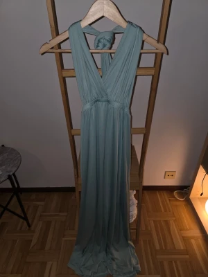 Ljusblå långklänning med omlott - Snygg ljusblå långklänning med omlottdesign och breda axelband som kan knytas på många olika sätt. Klänningen har ett mjukt, fallande tyg och markerad midja för en smickrande siluett. Perfekt för dig som vill ha en elegant och enkel look.