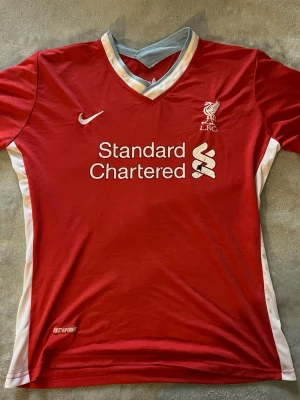 Liverpool Nike matchtröja röd - Liverpool FC matchtröja i rött med vita detaljer och V-ringning. Nike-logga och klubbmärke på bröstet samt sponsortryck 'Standard Chartered' framtill. Tillverkad i lätt och ventilerande funktionsmaterial, perfekt för fotbollsträning.