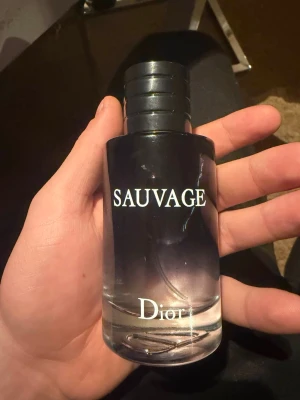 Dior Sauvage parfymflaska - Snygg parfymflaska från Dior, modellen Sauvage. Flaskan är cylindrisk med svart och transparent glas som tonar ut mot botten. Lock i svart plast med Dior-loggan på toppen. Perfekt för dig som gillar exklusiva dofter och stilren design.