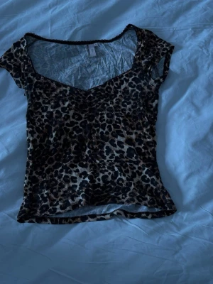 Leopardmönstrad topp från Nelly - Trendig leopardmönstrad topp från Nelly med v-ringning och korta ärmar. Tunn och lätt i materialet, perfekt för att skapa en edgy look. Svarta spetsdetaljer längs halsringningen ger en extra touch.