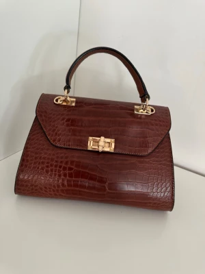 Brun krokopräglad handväska - Snygg handväska i mörkbrunt krokopräglat konstläder med guldfärgade metalldetaljer och vridlås. Väskan har ett stadigt handtag och elegant, klassisk form. Perfekt accessoar för dig som gillar tidlös och chic stil.