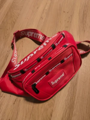 Röd Supreme bröst väska - Säljer en röd bröst väska från Supreme med tydliga loggor på både rem och framficka. Väskan har 4  fack med dragkedja och justerbar rem. Perfekt för att bära det viktigaste och ge outfiten en streetwear vibe.
