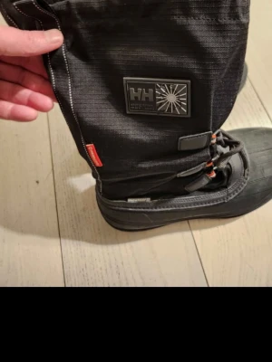 Svarta vinterstövlar från Helly Hansen - Säljer ett par svarta vinterstövlar från Helly Hansen med högt skaft, grov sula och varm, mjuk insida. Stövlarna har snörning framtill och detaljer i rött samt en tydlig HH-logga på sidan. Perfekta för kalla dagar och snöiga äventyr.