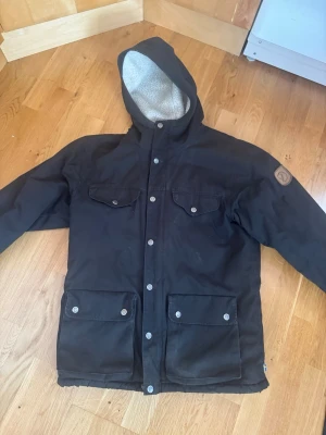 Svart parkajacka från Fjällräven XS Greenland - Svart parkajacka från Fjällräven med mysigt grått teddyfoder och huva. Jackan har fyra fickor med knappar framtill och stängs med både dragkedja och tryckknappar. Perfekt för kalla dagar och har en klassisk outdoor-look.