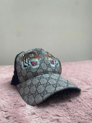 Zara keps med tigertryck och GG-mönster - Säljer en snygg Zara  keps med klassiskt GG-mönster i grått och svart. Framtill finns två detaljerade tigerhuvuden i färg. Kepsen har böjd skärm och mesh på baksidan för extra ventilation. Perfekt accessoar för dig som vill sticka ut.