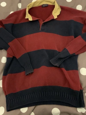 Randig stickad tröja från Gant - Snygg stickad tröja från Gant med breda marinblå och vinröda ränder samt en kontrasterande gul krage. Tröjan har långa ärmar, ribbstickade muddar och en klassisk rugbytröje-look. Perfekt för dig som gillar preppy stil. Storlek XXL
