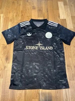 AJAX X STONE ISLAND - Snygg svart Ajax fotbollströja och Stone Island med diskret mönster, vita detaljer och klassiska Adidas-ränder på axlarna. Tröjan har krage, knapp framtill och loggor på bröstet. Perfekt för dig som gillar streetwear och sportig stil.