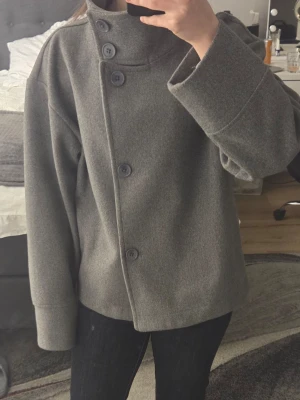 Grå ullrock med asymmetrisk knäppning - Snygg grå rock i ullmix med oversized passform och hög krage. Rocken har stora knappar i en asymmetrisk rad framtill och breda ärmar. Perfekt för dig som gillar minimalistisk och modern stil.