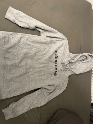 Grå hoodie från Björn Borg - Snygg grå hoodie från Björn Borg med svart logga tryckt på bröstet. Klassisk modell med huva och dragsnören, samt mjukt material som känns skönt mot huden. Perfekt för en avslappnad och sportig stil.