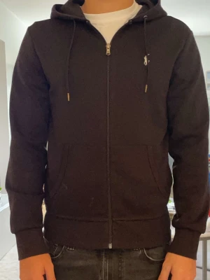 Svart hoodie med dragkedja Ralph Lauren - Svart hoodie med dragkedja från Ralph Lauren. Klassisk modell med huva och dragsko, två stora fickor framtill och liten vit logga på bröstet. Tillverkad i mjukt bomullsmaterial för skön känsla och enkel stil.
