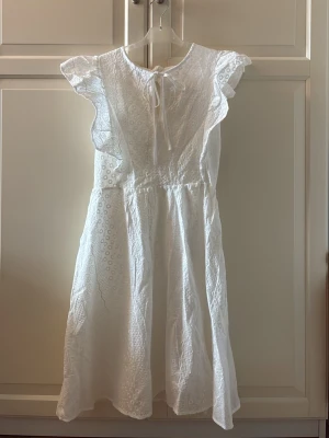 Vit spetsklänning med volangärm - Söt vit klänning med broderad spets, volangärmar och knytband i nacken. Klänningen har ett romantiskt mönster och är tillverkad i ett lätt material. Midjan är markerad och kjolen har ett fint fall. Öppen rygg. Storlek M. Helt ny och etiketten sitter kvar. 