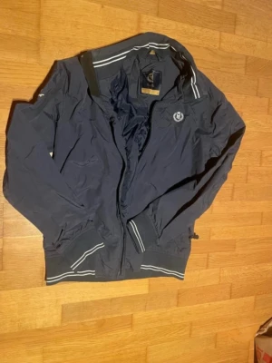 Marinblå vindjacka från Henri Lloyd - Snygg marinblå vindjacka från Henri Lloyd med vita ränder på krage och muddar. Superfintskick!!