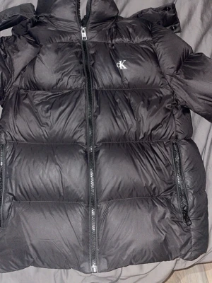 Svart pufferjacka från Calvin Klein - Snygg svart pufferjacka från Calvin Klein med diskret CK-logga på bröstet. Jackan har dragkedja framtill, två stora fickor med dragkedja och hög krage. Tillverkad i glansigt material som ger en modern look och håller dig varm under kalla dagar. Obs lite fläckig av smink men de går garanterat bort vid lite ansträngning 