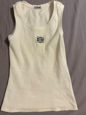 Vit ribbad linnetopp från Loewe - Säljer en vit ribbad linnetopp från Loewe med broderad logga i svart framtill. Toppen är ärmlös och har en klassisk rund halsringning. Perfekt att styla till jeans eller kjol för en clean och trendig look.