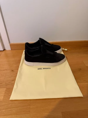 Svarta Axel Arigato - Säljer ett par svarta sneakers från Axel Arigato i mjuk mocka med vit sula. Klassisk låg modell utan snörning för clean look. Kommer med original dustbag. Perfekta för dig som gillar minimalistisk och stilren design.