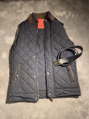 Svart quiltad dunväst Silver Stone M - Snygg svart quiltad dunväst från Silver Stone i storlek M. Västen har bruna detaljer vid fickorna och insidan av kragen, samt tryckknappar och dragkedja. Perfekt för lager-på-lager och ger en stilren look till din outfit.✅