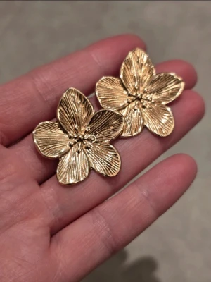Guldiga blomformade örhängen rostfritt stål - Säljer ett par stora, guldiga örhängen i form av blommor med fem kronblad. Örhängena har en detaljerad yta med fina linjer och små kulor i mitten som ger en lyxig och trendig känsla. Perfekta för dig som vill sticka ut med accessoarer i metallic.