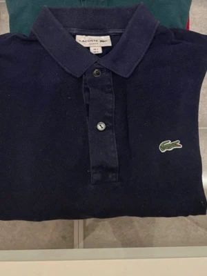 Mörkblå piké från Lacoste slim fit - Klassisk mörkblå pikétröja från Lacoste i slim fit-modell. Tröjan har krage, knappar framtill och den ikoniska gröna krokodilloggan på bröstet. Perfekt för en clean och stilren look.