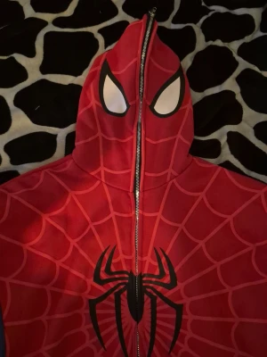 Spiderman hoodie med dragkedja - Röd och blå hoodie inspirerad av Spiderman med hel dragkedja framtill, vita ögon på huvan och svart spindeltryck på bröstet. Tjockt tyg med tryckt spindelnätsmönster över hela plagget.