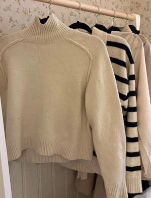 Vit polotröja i stickat material - Mysig vit stickad polotröja med lång ärm och ribbade muddar vid ärmslut och nederkant. Tröjan har en hög krage och en avslappnad passform, perfekt att matcha med jeans för en enkel och stilren look.