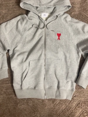 Grå zip hoodie från Ami Paris - Snygg ljusgrå hoodie från Ami Paris med dragkedja, huva med dragsko och känguruficka. På bröstet sitter Ami:s ikoniska röda hjärta med bokstaven A. Tillverkad i mjuk bomullsblandning som känns skön mot huden. Perfekt för en avslappnad och trendig look.