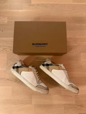 Burberry Sneakers - Snygga vita sneakers från Burberry med klassiskt rutmönster i beige, svart och rött på sidan. Skorna har detaljer i mocka och läder, samt vita snören och gummisula. Perfekta för dig som vill ha en stilren och lyxig look.