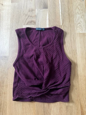 Vinröd ribbad croppad topp Bershka - Säljer en vinröd croppad topp från Bershka med twistad detalj framtill. Toppen är ribbad och ärmlös, perfekt för att styla med höga jeans eller kjol. Supersnygg färg och sitter tajt mot kroppen.