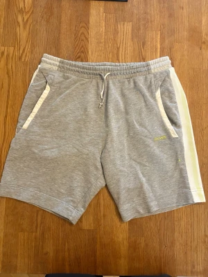 Grå shorts från BOSS med vita detaljer - Snygga grå shorts från BOSS med vita sidopaneler och gul logga på vänster ben. Shortsen har snörning i midjan och två fickor fram. Perfekta för en avslappnad och sportig stil.
