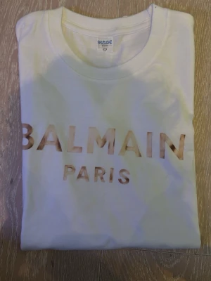 Vit Balmain Paris t-shirt - Snygg vit t-shirt från Balmain Paris med stort logotryck i guld på bröstet. Klassisk rund halsringning och korta ärmar. Perfekt för dig som gillar exklusiva streetwear-vibbar och vill sticka ut med ett ikoniskt märke. Samt 1 Moncler och en Emporio Armani 