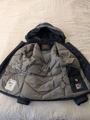 Mörkblå dunjacka från Woolrich - Snygg mörkblå dunjacka från Woolrich med avtagbar huva och stora fickor framtill. Jackan har dragkedja och knappar, samt quiltad insida i grått. Perfekt för kalla vinterdagar och riktigt varm tack vare dunfyllningen.