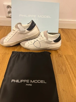 Vita Philippe Model sneakers - Snygga vita sneakers från Philippe Model med svarta detaljer på hälen och grå mocka vid tån. Skorna har en klassisk låg siluett, vita skosnören och diskret logga på sidan. Tillverkade i läder och mocka för en exklusiv känsla. Perfekta för en trendig streetstyle-look.