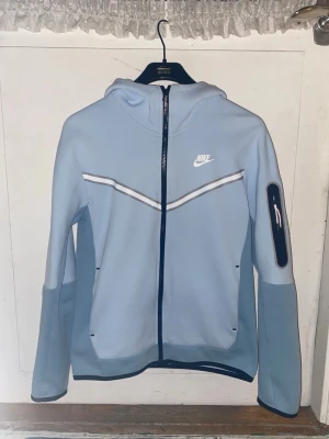 Blå Nike Tech Fleece Hoodie - En blå Nike Tech Fleece hoodie med vita och svarta detaljer i storlek S