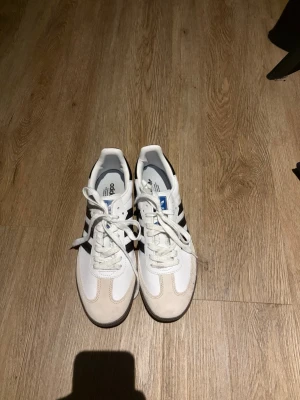 Adidas Samba vita och svarta sneakers - Klassiska Adidas Samba sneakers i vitt och svart med blå detalj på plösen. Ovandel i läder och mocka, med de ikoniska tre svarta ränderna på sidan. Yttersula i gummi med mönster för bra grepp. Perfekta för en sportig och stilren look.