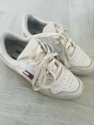 Vita Tommy Hilfiger sneakers - Snygga vita sneakers från Tommy Hilfiger med klassisk logga på sidan. Skorna har lågt skaft, perforerade detaljer på tån och ovandel i läder och textil. Perfekta för en clean och stilren look.