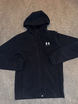 Svart vindjacka från Under Armour - Snygg svart vindjacka från Under Armour med huva och dragkedja framtill. Jackan har elastiska muddar vid ärmsluten, två sidofickor och vitt logotryck på bröstet. Tillverkad i lätt och vattenavvisande material, perfekt för blåsiga dagar. Och så du vet så är det en håll i den högra armen!!