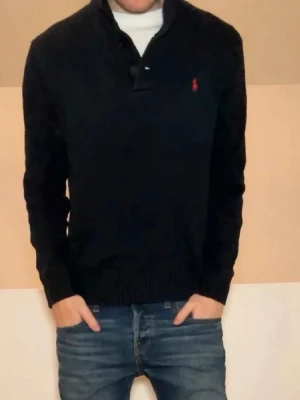 Svart tröja från Ralph Lauren - Klassisk svart tröja från Ralph Lauren med halv dragkedja och röd broderad logga på bröstet. Tröjan har ribbade muddar vid ärmslut och nederkant samt en hög krage. Perfekt till jeans för en stilren look.