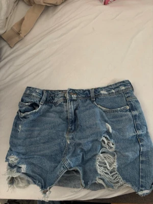 Blå slitna jeansshorts med fransar - Säljer ett par blå jeansshorts med slitna detaljer och fransar längst ner. Shortsen har klassisk femficksmodell, knappgylf och råa kanter. Perfekta för sommaren och ger en avslappnad vibe till din outfit.