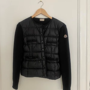 Svart cardiganjacka från Moncler - Snygg svart cardiganjacka från Moncler med quiltad front och stickade ärmar. Jackan har rund halsringning, knappstängning och fyra fickor fram. Moncler-logga på ena ärmen ger en exklusiv touch. Perfekt för lager på lager. Qr-kod och box. 